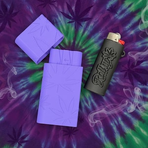 May include: A purple lighter case and a black lighter with the word "Rollup" and a cannabis leaf design. The case is open, revealing the lighter, and is decorated with cannabis leaves. The background is a tie-dye pattern.