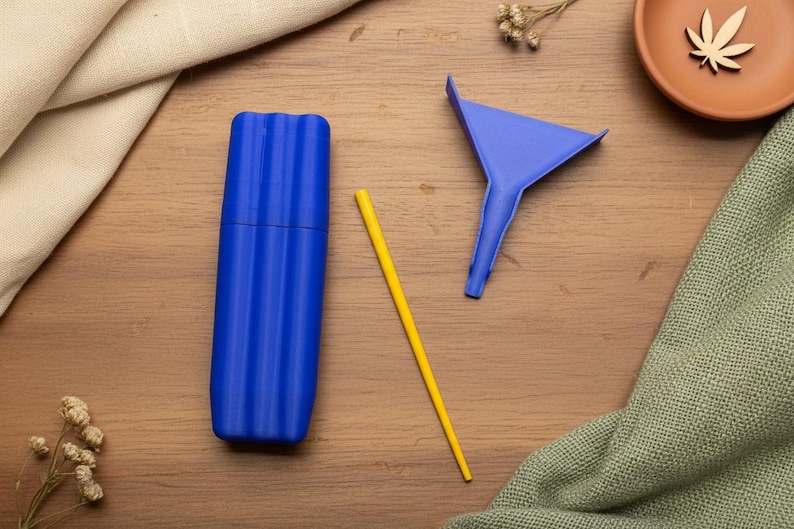 May include: A blue, ribbed, cylindrical container, a yellow straw, and a blue funnel are arranged on a wooden surface. A small terracotta dish with a wooden cannabis leaf is in the background.