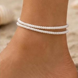 Puede incluir: Una tobillera de doble hilo con pequeñas cuentas blancas uniformes. La joya consta de dos bandas circulares finas y separadas que se llevan juntas alrededor del tobillo, mostrando una textura sencilla y un color blanco sólido y brillante.