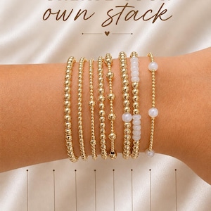 May include: A collection of seven gold-toned beaded bracelets featuring various bead sizes and textures, including some with white pearl-like accents. The text "CREATE YOUR own stack" appears at the top, with labels for each bracelet style below.
