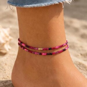 Könnte beinhalten: Ein zweireihiges Fußkettchen mit kleinen, runden Perlen in Magenta, Pink und Schwarz. Die obere Reihe enthält drei kleine, goldfarbene Metallperlen in der Mitte, die einen dezenten Kontrast zu dem lebendigen Muster aus pinken und schwarzen Perlen bilden.