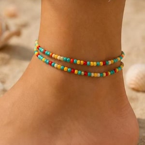 Puede incluir: Una tobillera de doble hebra con pequeñas cuentas redondas en un patrón repetitivo de turquesa, rojo, amarillo y blanco roto. Las dos hebras se usan juntas, creando un aspecto superpuesto con cuentas vibrantes y multicolores.