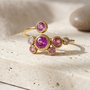 May include: A gold open ring featuring a cluster of round, bezel-set gemstones in varying shades of pink and clear white. The design includes one large central pink stone surrounded by smaller pink and white stones arranged in a circular pattern.