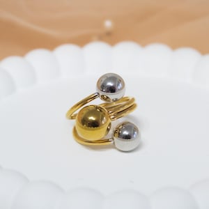 May include: A gold-coloured ring with a spiral design, featuring three spherical accents: one gold and two silver. The ring is displayed on a white, scalloped plate, showcasing its modern, minimalist style.