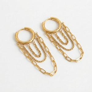 May include: A pair of gold-colored hoop earrings with multiple dangling chains of varying lengths and styles. The earrings have a polished finish and are displayed on a white background. The chains include small and large links.