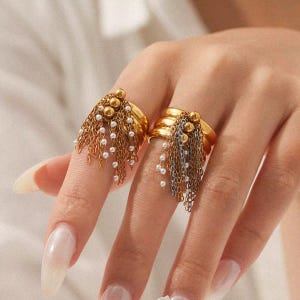 May include: Two gold rings with dangling chains and small white beads. One ring has gold chains and beads, the other has silver chains and beads. The rings are on a person's fingers.