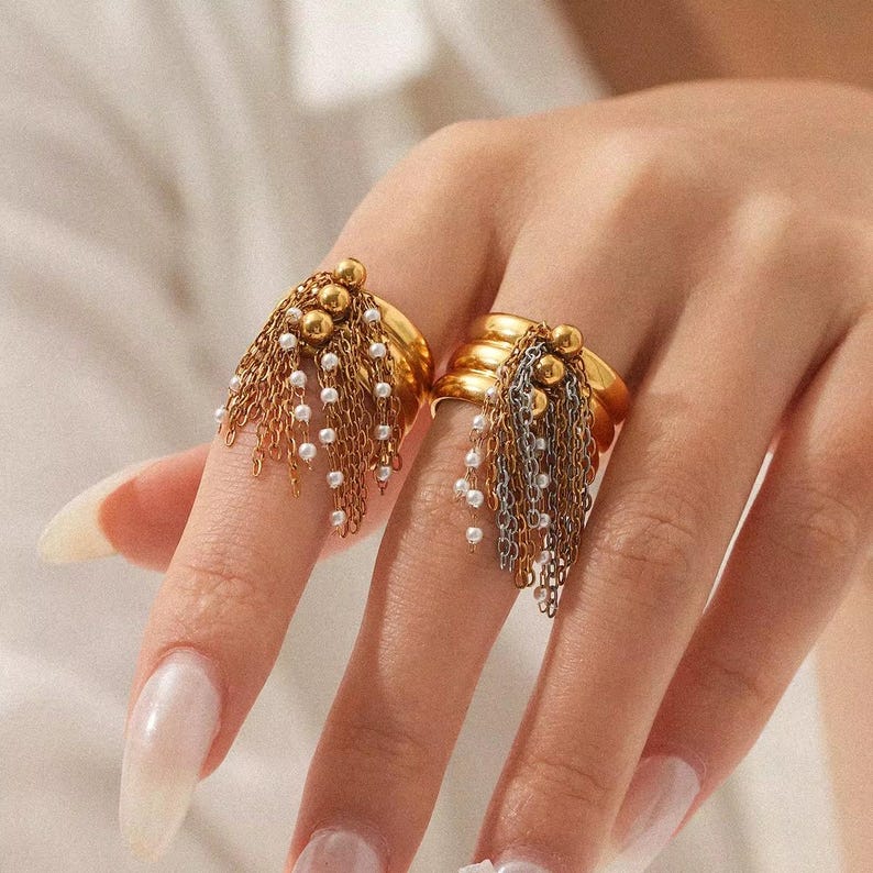 May include: Two gold rings with dangling chains and small white beads. One ring has gold chains and beads, the other has silver chains and beads. The rings are on a person's fingers.