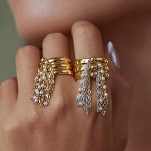 May include: Two gold rings with layered bands and dangling chains adorned with small white beads. One ring features gold chains, while the other combines gold and silver chains. The rings are worn on fingers.