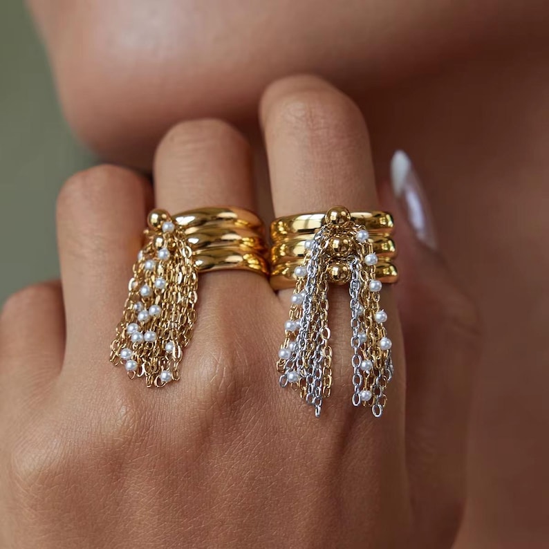 May include: Two gold rings with layered bands and dangling chains adorned with small white beads. One ring features gold chains, while the other combines gold and silver chains. The rings are worn on fingers.