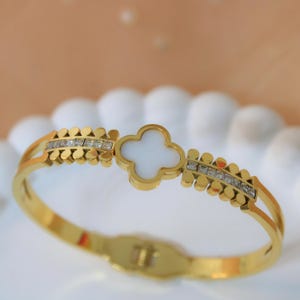 May include: Gold-coloured bracelet with a white clover-shaped centrepiece. The hinged bracelet features small, clear crystal accents and a textured pattern. The clasp is visible at the bottom of the bracelet.