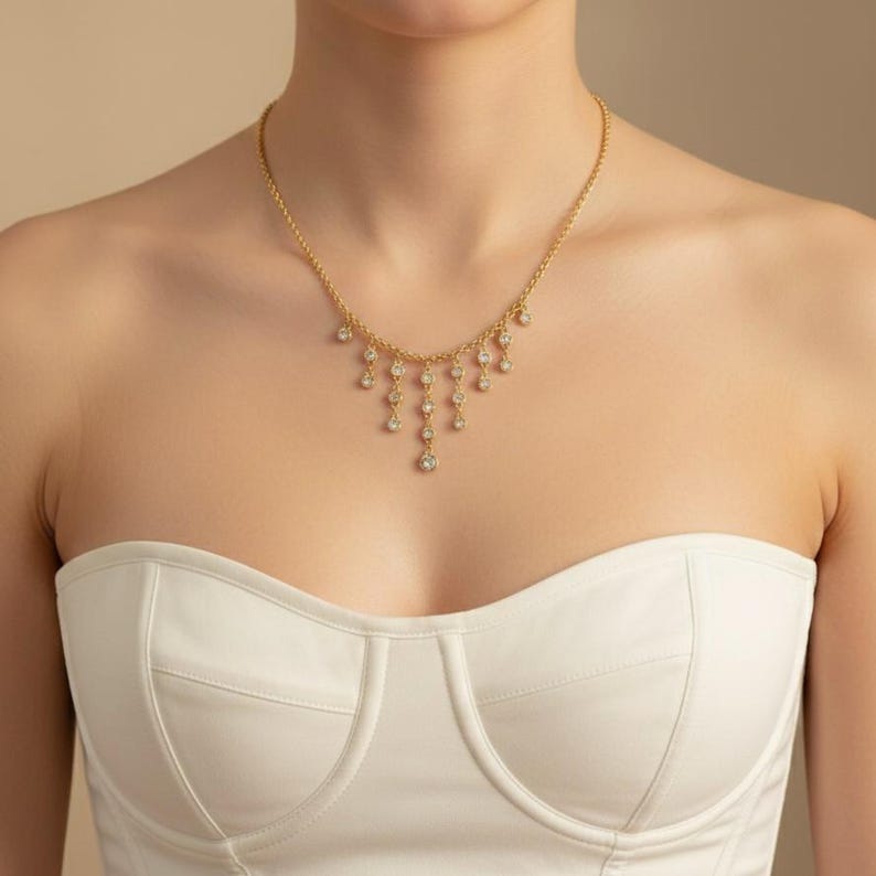 May include: A gold-toned necklace with a delicate chain and cascading diamond-like pendants. The necklace is worn against a white strapless top. The pendants are arranged in a graduated, teardrop design, adding a touch of elegance.