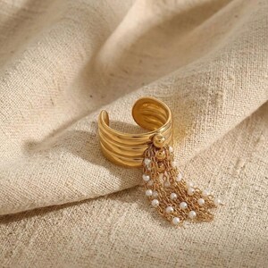 High-fashion editorial shot of a gold tassel ring resting on draped neutral fabric. The light highlights the polished 18k gold finish and the elegant drape of the dangling pearl chains.
