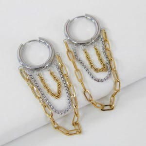 May include: A pair of silver hoop earrings with dangling chains in gold and silver tones. The earrings feature a mix of chain styles, including small and large links, creating a layered effect. The jewelry is displayed on a white surface.