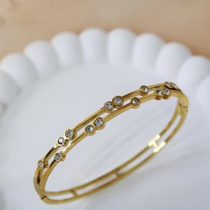 Peut inclure: Un bracelet jonc doré avec deux bandes parallèles. Les bandes sont ornées de petits pierres précieuses rondes et transparentes. Le bracelet est posé sur un fond blanc festonné, ce qui met en valeur le design du bijou.