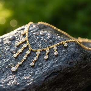 May include: A gold-colored necklace with multiple dangling diamond-like pendants. The necklace is displayed on a dark gray, wet rock with a blurred green background. The pendants are round and evenly spaced.