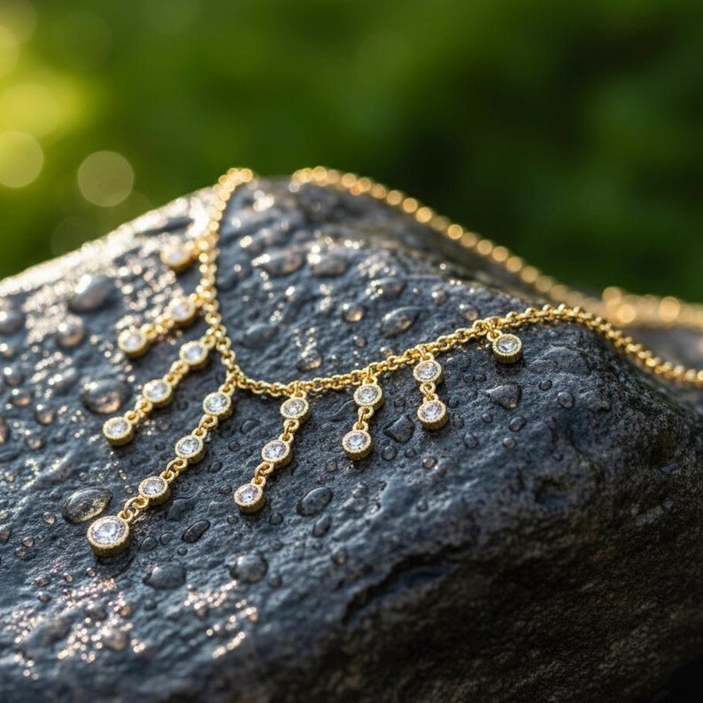 May include: A gold-colored necklace with multiple dangling diamond-like pendants. The necklace is displayed on a dark gray, wet rock with a blurred green background. The pendants are round and evenly spaced.
