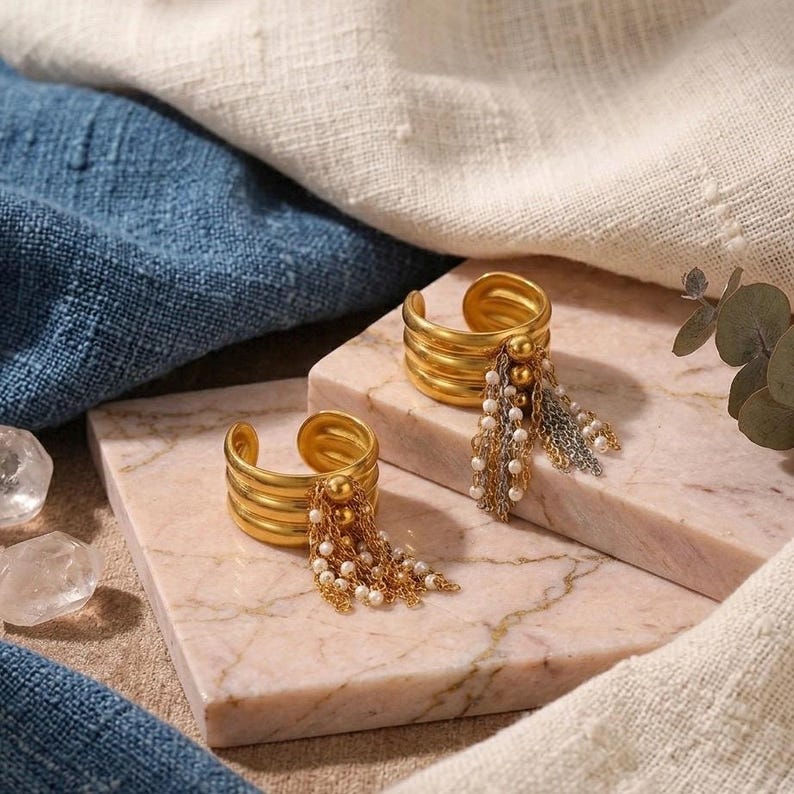 May include: Two gold-colored cuff rings with a layered design and dangling beaded and chain accents. The rings are displayed on a pink marble surface, with blue and white fabric in the background. The jewelry is a fashion accessory.