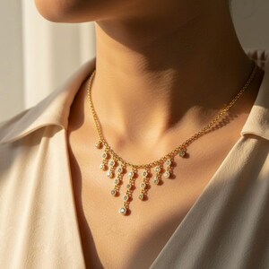 May include: A gold-colored necklace with a delicate chain and multiple dangling, round, clear gemstones. The necklace is worn around the neck, with the gemstones cascading down. The background is a neutral beige.