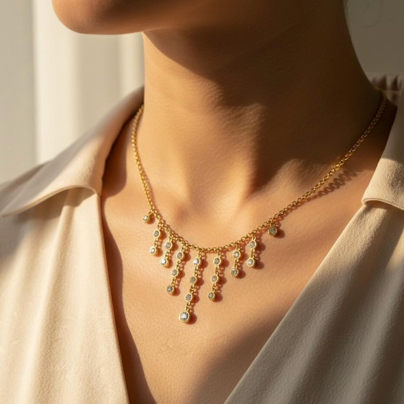 May include: A gold-colored necklace with a delicate chain and multiple dangling, round, clear gemstones. The necklace is worn around the neck, with the gemstones cascading down. The background is a neutral beige.