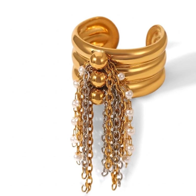 May include: A gold-toned cuff bracelet with a layered design. The bracelet features dangling chains with small pearls and gold and silver-toned chains. The cuff has a rounded shape and a smooth, polished finish.