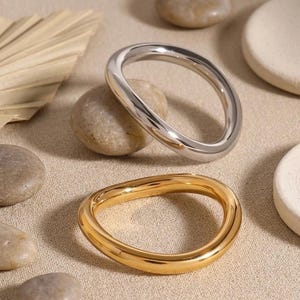 May include: Two smooth, irregular, organic-shaped rings, one in a polished silver-tone metal and one in a polished gold-tone metal. Both rings feature a continuous, rounded band with a fluid, non-circular silhouette and a high-shine reflective finish.