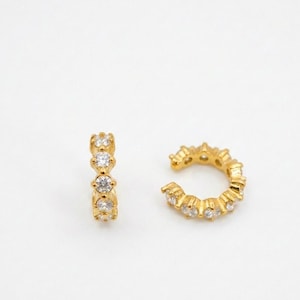 May include: Two gold-colored ear cuffs with clear, round gemstones. One cuff is a straight bar with four gemstones. The other is a curved ear cuff with six gemstones. The jewelry is set against a white background.