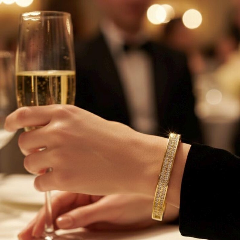 May include: A gold-colored bracelet with two rows of sparkling stones is worn on a wrist. A person holds a champagne flute. The background is blurred, suggesting a formal event or celebration.