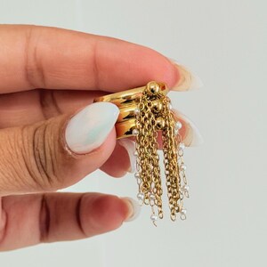 May include: Gold-toned rings with dangling chains of small pearls. The rings are stacked and held in a hand with manicured nails. The chains hang down, creating a layered effect. The jewelry is set against a plain background.