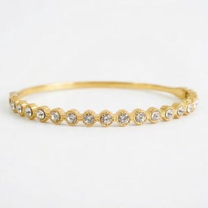 May include: A gold-colored bangle bracelet with a row of clear, round gemstones. The gemstones are evenly spaced around the bracelet, which has a simple, elegant design. The bracelet is set against a white background.