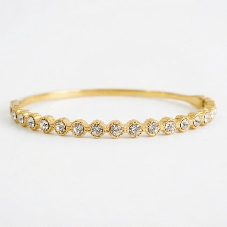 Puede incluir: Una pulsera de brazalete dorada con una fila de piedras preciosas redondas y transparentes. Las piedras preciosas est&aacute;n espaciadas uniformemente alrededor de la pulsera, que tiene un dise&ntilde;o simple y elegante. La pulsera est&aacute; sobre un fondo blanco.
