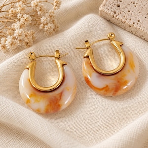 May include: A pair of gold-tone hoop earrings featuring a thick, crescent-shaped resin attachment with a marbled pattern in shades of white, cream, and amber. The earrings have a polished metallic finish and a secure hinged closure mechanism.