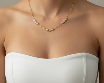 May include: A gold-toned necklace with a delicate chain and a series of clear, round gemstones. The gemstones are evenly spaced along the necklace, creating a subtle sparkle. The necklace is worn against a white strapless top.