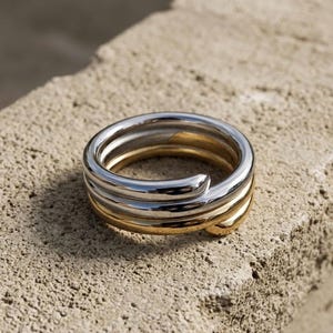 May include: A silver and gold ring with a layered design. The ring features three bands, two silver and one gold. The ring is set against a textured, light brown surface.