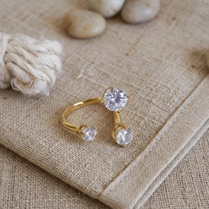 May include: A gold ring featuring three clear, round gemstones. The ring is displayed on a tan linen surface, with a small ball of white string and a few small, smooth stones in the background. The ring's design has two gemstones on either side of the finger and one on top.