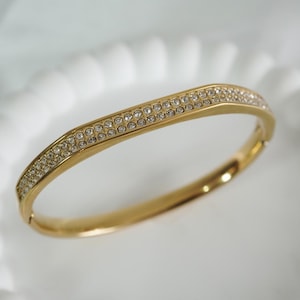 May include: A gold-toned bangle bracelet with two rows of small, clear crystals. The bracelet has a hinged clasp and a slightly curved design. The crystals sparkle against the gold metal, creating a glamorous look. This piece of jewelry is suitable for special occasions.