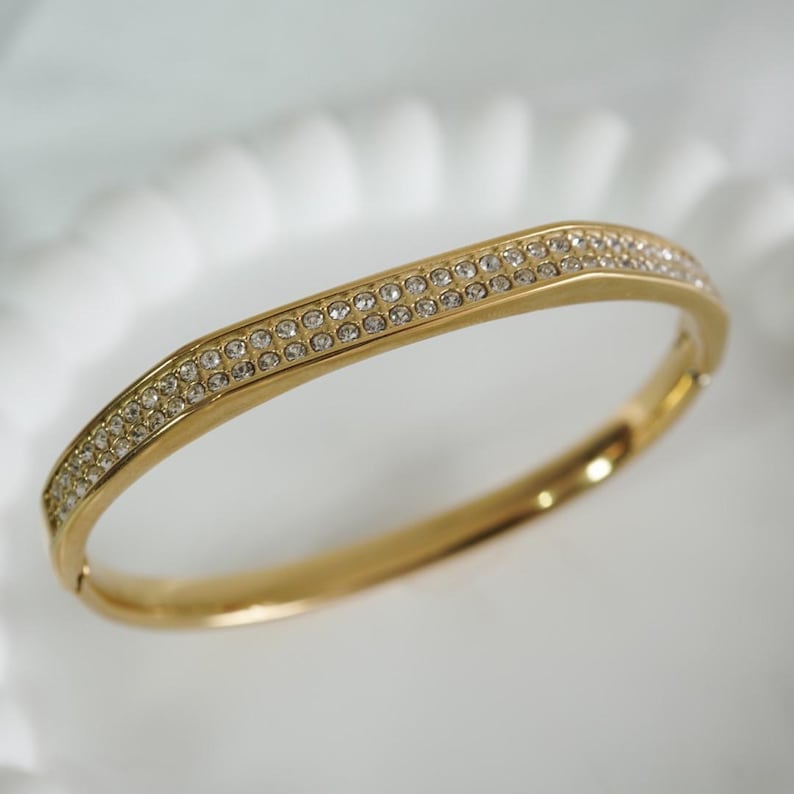 May include: A gold-toned bangle bracelet with two rows of small, clear crystals. The bracelet has a hinged clasp and a slightly curved design. The crystals sparkle against the gold metal, creating a glamorous look. This piece of jewelry is suitable for special occasions.