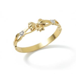 Puede incluir: Pulsera dorada con un diseño de lazo en el centro. La pulsera tiene un diseño delicado y calado con detalles en forma de corazón y redondos, y pequeñas piedras preciosas transparentes. El estilo general es elegante y femenino.