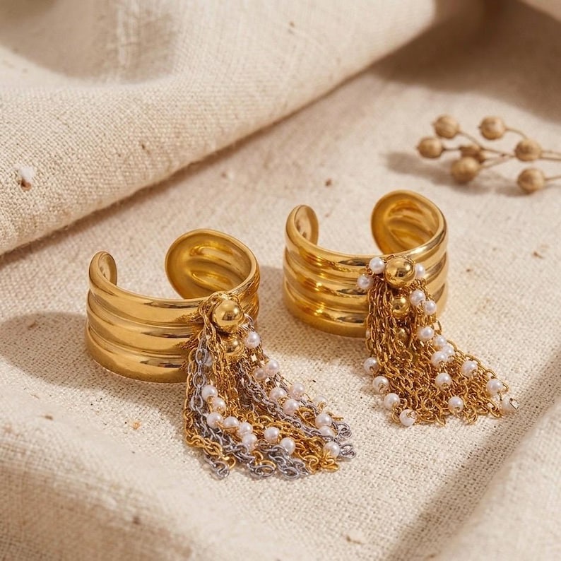 May include: Two gold-colored cuff rings with layered bands and dangling chains. Each ring features a cluster of small white pearls and mixed gold and silver chains. The rings are displayed on a neutral-colored fabric.