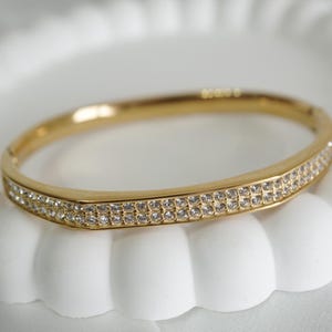 May include: A gold-toned bangle bracelet with two rows of small, clear crystals. The bracelet has a hinged clasp and a smooth, polished finish. The crystals sparkle, adding a touch of elegance to the jewelry.