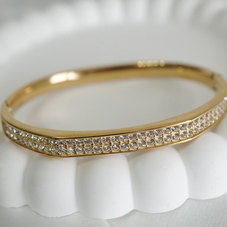 May include: A gold-toned bangle bracelet with two rows of small, clear crystals. The bracelet has a hinged clasp and a smooth, polished finish. The crystals sparkle, adding a touch of elegance to the jewelry.