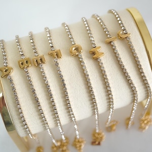 May include: Gold-coloured initial bracelets with a delicate rhinestone chain. Each bracelet features a different letter, displayed on a white surface. The bracelets are displayed on a gold-coloured stand.