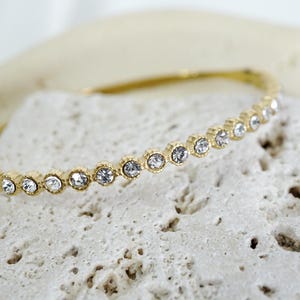 Puede incluir: Una pulsera dorada con una fila de cristales claros y brillantes. La pulsera se encuentra sobre una superficie texturizada de color blanquecino, creando un contraste entre el metal liso y el fondo rugoso. Joyer&iacute;a.