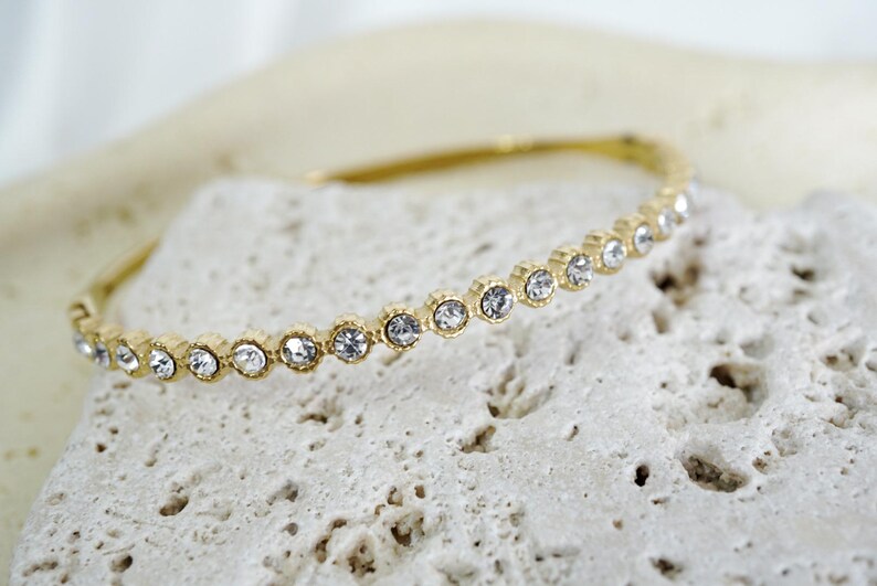Puede incluir: Una pulsera dorada con una fila de cristales claros y brillantes. La pulsera se encuentra sobre una superficie texturizada de color blanquecino, creando un contraste entre el metal liso y el fondo rugoso. Joyer&iacute;a.
