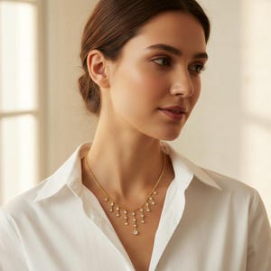 May include: A gold necklace with a delicate chain and multiple dangling, round, clear gemstones. The necklace is worn against a white button-down shirt. The gemstones are arranged in a cascading pattern, with a larger stone at the bottom.