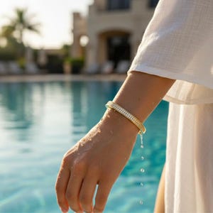 May include: A gold-colored bangle bracelet with two rows of small, sparkling stones. The bracelet is worn on a wrist with water droplets, suggesting a poolside or beach setting. The bracelet is a piece of jewelry.