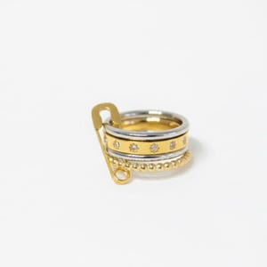 May include: A collection of four rings in gold and silver hues, secured by a gold safety pin. The rings feature a beaded band, a band with small clear stones, and two bands with a gold finish.