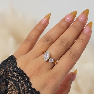 May include: A gold ring featuring a marquise-cut diamond and a smaller round diamond. The ring is set on a slender gold band. The hand is adorned with long nails with a gradient design, transitioning from pink to gold.