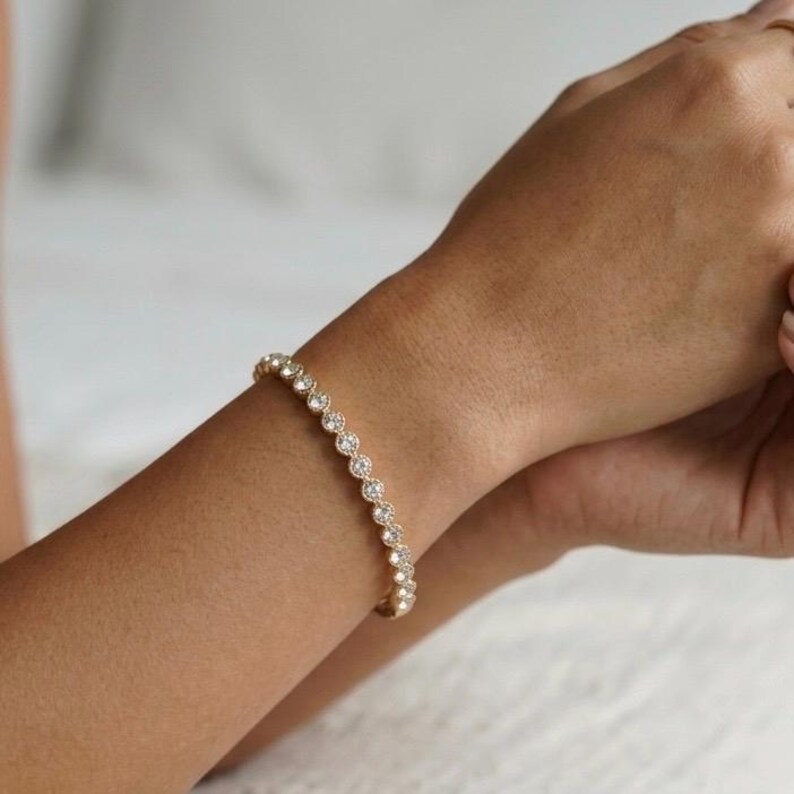 Puede incluir: Un brazalete dorado con una serie de peque&ntilde;as piedras preciosas redondas y transparentes. El brazalete se lleva en la mu&ntilde;eca, mostrando su delicado dise&ntilde;o y brillo. Las piedras est&aacute;n espaciadas uniformemente.