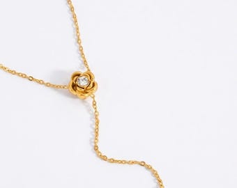 May include: Gold-colored necklace featuring a floral pendant with a central clear gemstone. The necklace has a delicate chain design, with a second, smaller floral pendant at the end of a dangling chain. The background is plain white.