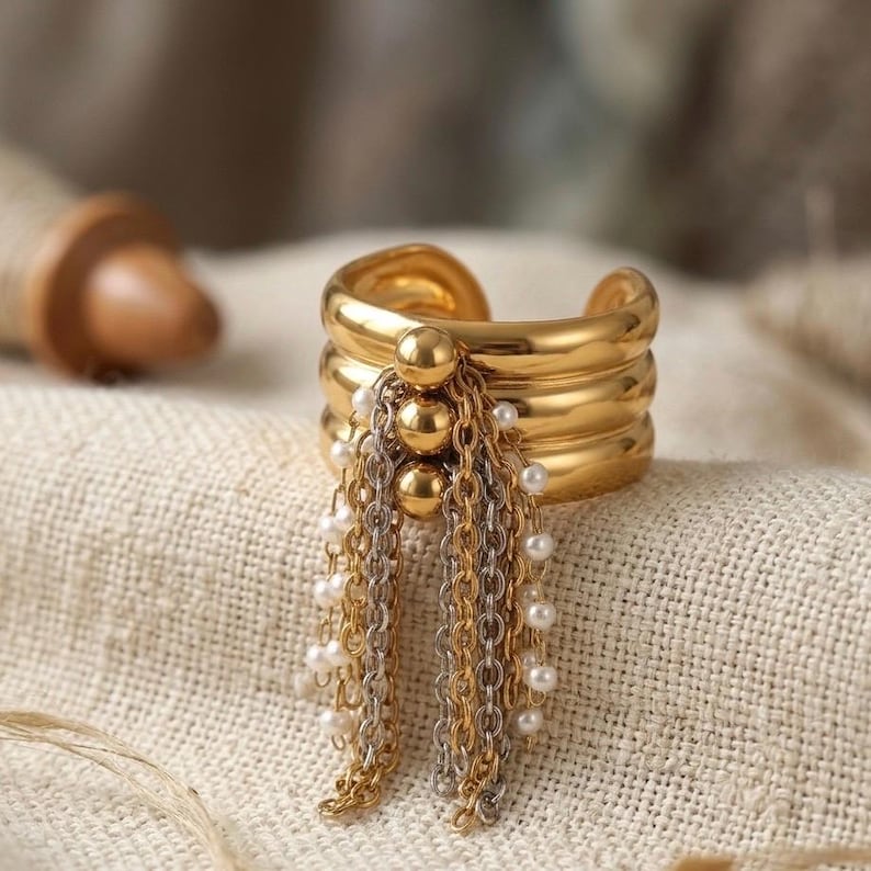 Close-up of a luxury gold triple-band cuff ring on a beige linen surface. A cluster of three gold beads holds a cascading tassel of gold and silver chains interspersed with small white seed pearls.